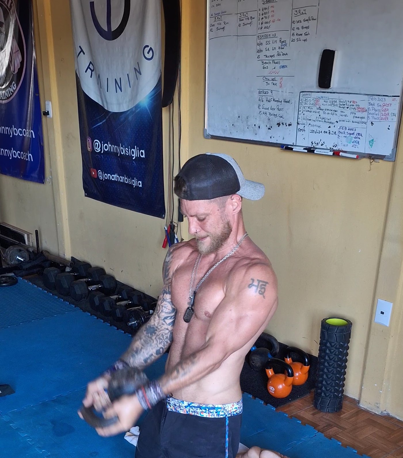 El coach Johnny entrenando con kettlebells sobre las colchonetas azules del estudio, con los banners de JB Training y la pizarra de rutinas atrás